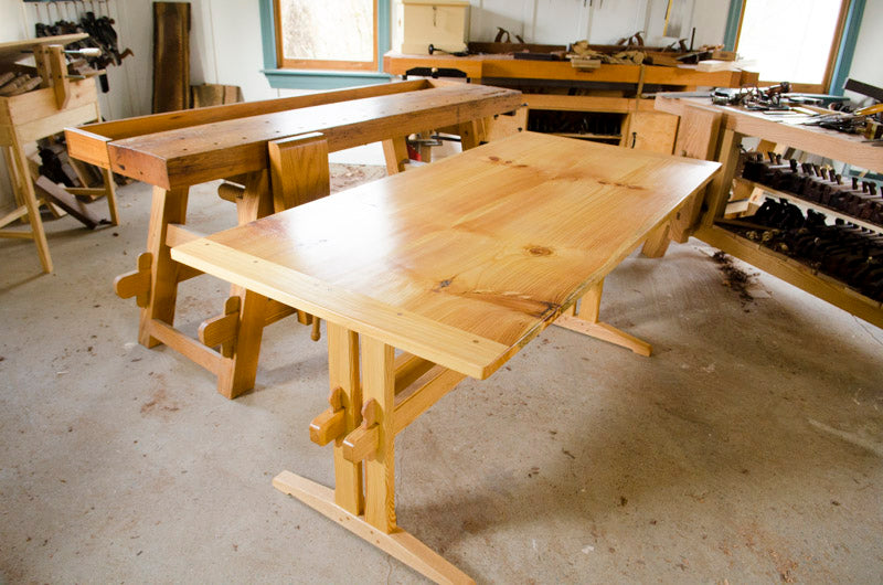 Finished trestle table sitting next to woodworking workbenches in Joshua Farnsworth's woodworking workshop